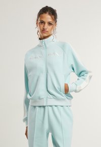 Šviesiai mėlynas zip-up treniruočių švarkas su ribotu manžetų dizainu, šoninėmis kišenėmis ir baltomis detalėmis; turi iškilų "KARL KANI" logotipą ant krūtinės.
