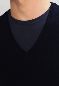 Mörkblå V-ringad tröja i mjukt material, lager på lager över en mörkblå skjorta. Texturen ser slät ut med ribbade kanter.