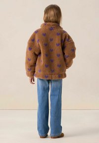 Veste en polaire marron avec des motifs de cœurs violets, col montant et coupe décontractée. Associée avec un jean évasé bleu clair et des chaussures marron.