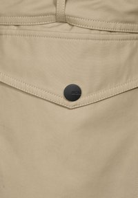 Beiger Stoff mit glatter Textur besitzt eine dreieckige Tasche, die von kontrastierenden Nähten umrissen ist, sowie einem schwarzen Knopf mit eingraviertem "CECIL".