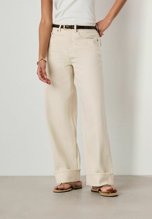 Person wearing high-waisted wide-leg beige pants with rolled cuffs, black belt, white sleeveless top, gold bracelet, and brown sandals, standing.