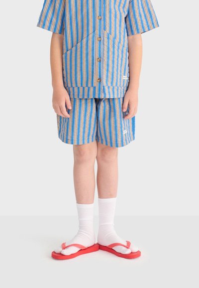 Enfant portant une chemise à manches courtes rayée bleu et beige et un short assorti, chaussettes blanches et tongs rouges, debout devant un fond neutre.