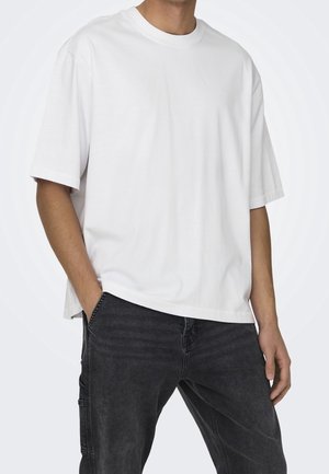 Person iført en ensfarvet hvid kortærmet oversized T-shirt og sorte jeans med den ene hånd i lommen mod en ensfarvet baggrund.