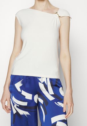 Witte, getailleerde top met een asymmetrische halslijn en een geplooide schouderdetail, voorzien van een gouden accent. Draag het met een patroon in blauwe broek.