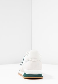 Baskets en daim blanc avec accents verts, comportant un talon texturé, un devant à lacets et une semelle en caoutchouc marron. Logo Lacoste sur le talon.