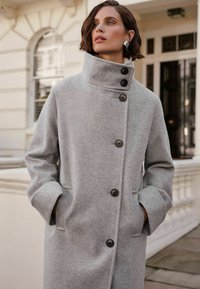 Cappotto di lana grigio con colletto alto, grandi bottoni frontali e tasche laterali; presenta una texture morbida e una vestibilità rilassata.