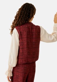 Chaleco rojo con un diseño floral texturizado. Camisa de manga larga color crema con puños fruncidos. Vista desde la parte trasera.