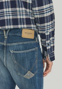 Jeans in denim blu con un'etichetta visibile con la scritta "ROY ROGER'S". Presentano un motivo di cucitura diagonale sulla tasca posteriore e un tessuto testurizzato.