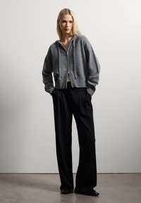 Felpa grigia in lana con zip frontale, design corto e orlo a costine, abbinata a pantaloni neri a gamba larga, esposta su uno sfondo neutro.
