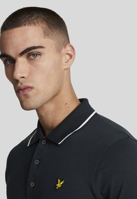 Svart polo skjorte med hvit kragepiping, tre knapper og en gul brodert logo på venstre bryst. Glatt bomullstekstur.