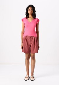 Haut rose côtelé avec des manches courtes à volants, associé à un short texturé couleur rouille taille haute. Le mannequin porte des chaussures plates de couleur claire.
