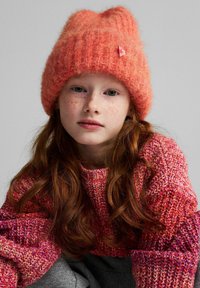 Pomarańczowa dziergana czapka z mankietem i małym różowym logo; noszona z teksturowanym różowo-czerwonym swetrem. Widoczne długie czerwone włosy.