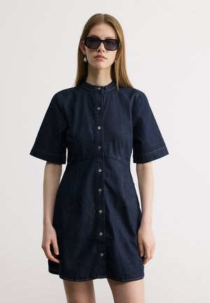 Mujer con vestido de mezclilla azul oscuro abotonado, de manga corta, y grandes gafas de sol negras, de pie frente a un fondo claro y sencillo.