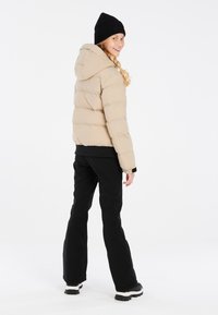 Beige Daunenjacke mit Kapuze, gerippte schwarze Mütze, schwarze Hose und robuste schwarze Stiefel. Glatte Textur mit gestepptem Design.