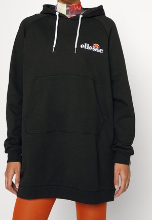 Czarna bluza typu oversized z białymi sznurkami i logo Ellesse na piersi, noszona na kolorowym golfie i pomarańczowych spodniach.