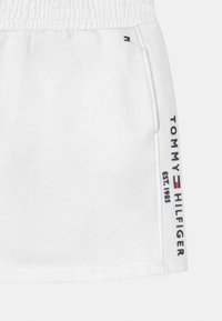 Vita shorts med elastisk midja, med "TOMMY HILFIGER" tryckt i svart längs sidan, och en liten logotyp i röd, vit och blå.