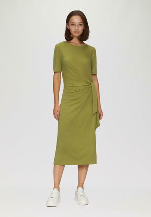 Olivgrünes, knielanges Kleid mit kurzen Ärmeln und einer gedrehten Vorderseite. Kombiniert mit weißen Plateausneakern für einen Kontrast. Glatte Textur.