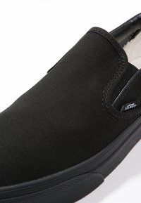 Chaussure Vans noire slip-on avec accents élastiques sur les côtés et semelle en caoutchouc texturée, avec une petite étiquette logo Vans près du talon.