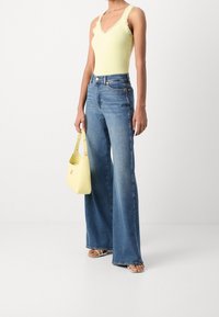 Kvinna som bär en ljusgul ärmlös topp med spetsdetaljer, högmidjade vida blå jeans, vita sandaler och håller en gul handväska.