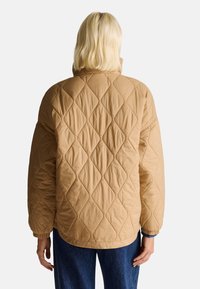 Gepolsterte, übergroße, beige Jacke mit hohem Kragen, die ein Rautenmuster und elastische Bündchen aufweist. Getragen über Jeanshosen.