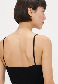 Haut en maille noire à fines bretelles, présentant un tissu texturé et une bordure festonnée le long des bords, vu de dos.