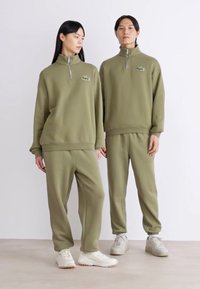 Sudadera con cremallera de color verde oliva y pantalones a juego, hechos de tela suave. Presenta un pequeño logo bordado en el pecho. Diseño casual.