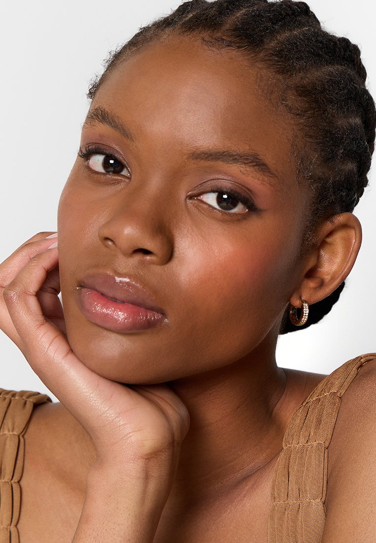 Primer plano de una persona con piel oscura, que lleva pequeños pendientes de aro de oro y una blusa de textura bronceada, sosteniéndose la cara con una mano.