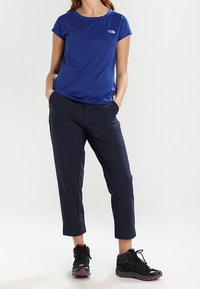 Sininen lyhythihainen urheilupaita, jossa on logo, yhdistettynä tummansinisiin kevyisiin housuihin ja mustiin vaelluskengiin, joissa on purppuran värisiä yksityiskohtia.