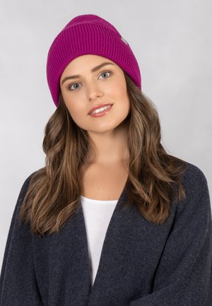 Fuchsia ribbed knit beanie with a snug fit, featuring a small logo tag. Model wears a white top and dark blue cardigan, with wavy hair.
