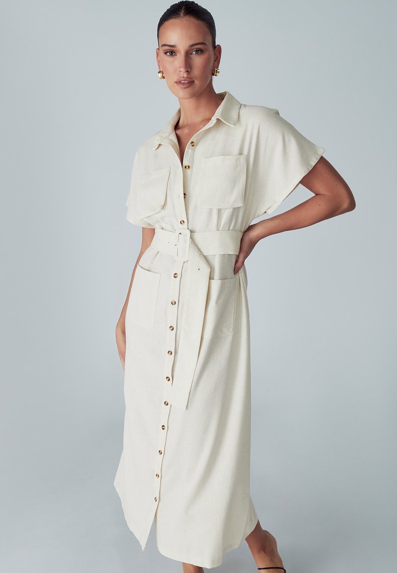 Beige Leinenhemdkleid mit kurzen Ärmeln, Knopfleiste, zwei Brusttaschen, einem Stoffgürtel und einem schulterhohen Schlitz am unteren Ende.