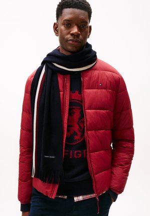 Giacca a vento rossa con design trapuntato, indossata sopra un maglione blu navy con una grafica di un leone rosso. Abbinata a una sciarpa a righe blu navy e bianche.