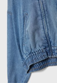 Veste en denim bleu clair en gros plan montrant une ceinture élastique, une fermeture éclair et une poche latérale verticale avec des détails de couture visibles.