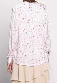 Blouse légère à motifs floraux, manches longues, coupe ample et ourlet arrondi, fabriquée en tissu lisse blanc avec des accents rouges et roses.