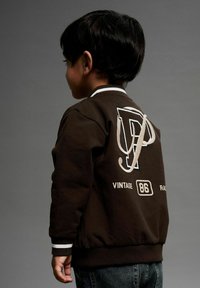 Bruine bomberjack met witte accenten, met op de achterkant een groot logo met de letter "P" en de tekst "VINTAGE 86 RACING" eronder.