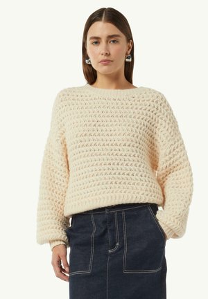 Cream-colored knit sweater featuring a textured, loose weave design with ribbed cuffs and hem. Paired with a dark denim skirt.