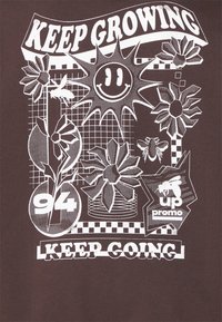 Brun t-skjorte med hvitt grafisk design med blomster, en smilende sol, og tekstene: "KEEP GROWING," "94," og "KEEP GOING." Mønstrene inkluderer rutenett og sjakkmønster.