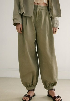 Luźne oliwkowo-zielone spodnie z elastycznymi mankietami, noszone z brązowymi sandałami na sprzączkę, krótką kurtką i złotymi pierścionkami na obu dłoniach.