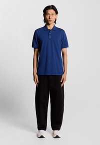 Blå polo med slät tyg och krage, prydd med en liten broderad logotyp på bröstet, tillsammans med svarta vida byxor och sneakers.