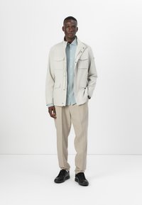 Light beige jacket with four pockets, layered over a light blue shirt, paired with beige trousers and black sneakers. Simple and functional design.