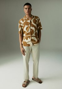 Kortebloemshirt in bruine en crèmekleurige abstracte patronen, gecombineerd met lichtbeige broek en grijze sandalen, op een neutrale achtergrond.
