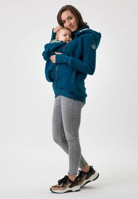 Felpa con cappuccio color turchese e tasca frontale per bambino, abbinata a jeans skinny grigi chiari e sneakers nere con accenti multicolori.