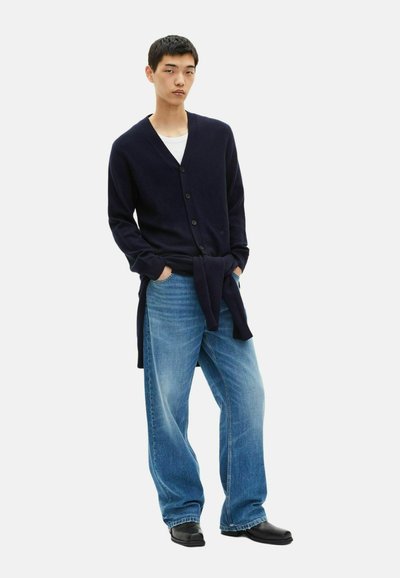Cardigan marine avec un col en V et fermeture à boutons, associé à un jean bleu clair à jambes larges et des chaussures noires. Design simple, texture douce.