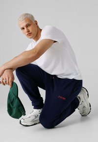 Bela majica s kratkimi rokavi in temno modre športne hlače, z majhnim rdečim logotipom. Drži zeleno kape, obuta v bele superge s črnimi poudarki.