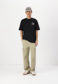 Sort bomulds t-shirt med hvid tekst på brystet, khaki cargo bukser med sidelommer, og grå kasket. Fodtøj: hvide sneakers med brune såler.
