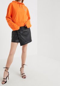 Haut de survêtement court orange avec une poche avant et un cordon de serrage, associé à une mini-jupe en cuir noire avec un design à enroulement sur le côté et des talons noirs à lanières.