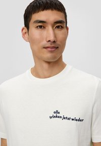 Valkoinen puuvillainen t-paita, jossa on pyöreä kaula-aukko ja lyhyet hihat, ja jonka rinnassa on tummansininen teksti "alle trinken jetzt wieder".