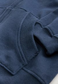 Nærbillede af en marineblå sweatshirtlomme med ribbet tekstur på ærmerne og lommens kant.