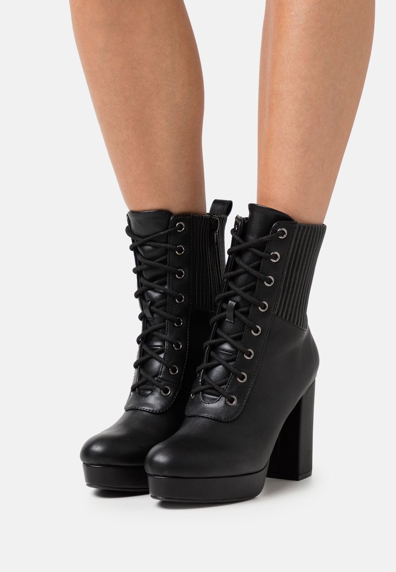 Bottines noires en cuir à lacets avec talons hauts épais et détail en tissu côtelé à l'arrière, portées sur des jambes nues sur un fond uni.
