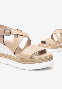 Sandali in pelle beige con cinturini incrociati, ampia apertura per le dita dei piedi e suola piatta con un accento in juta intrecciata e base in gomma bianca.