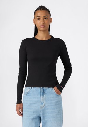 Mujer con el cabello trenzado que lleva una camiseta negra ajustada de manga larga y vaqueros azul claro con una mano en el bolsillo, sobre un fondo blanco.